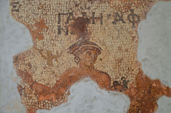Mosaic of Aphrodite. Most of her body is missing but we see her shoulders, her right arm with a bracelet and her head, adorned with a crown. She also wears earrings. It's notable that this depiction of Aphrodite is fairly tan compared to the pale tones commonly used for women in Graeco-Roman art. A fragment of her name remains in Greek letters above her.