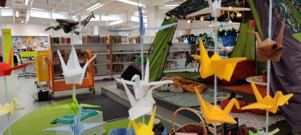Interior of a local public library in Iso Omena mall in Espoo, Finland. Yellow, white, red and blue paper origami birds hanging from the ceiling in front of a green story telling and reading tent. Background space has low bookshelves.