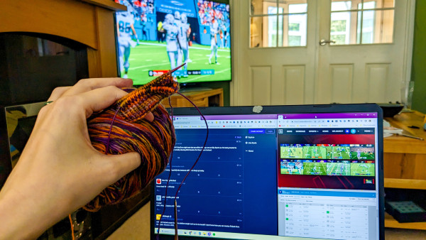 My hand holds the beginning few rounds of sock ribbing and a yarn cake in oranges and purples. Behind that, my laptop screen with NFL RedZone, fantasy, and the NFL hashtag feed on Mastodon. Behind THAT, our TV is showing tonight's Miami Dolphins game.
