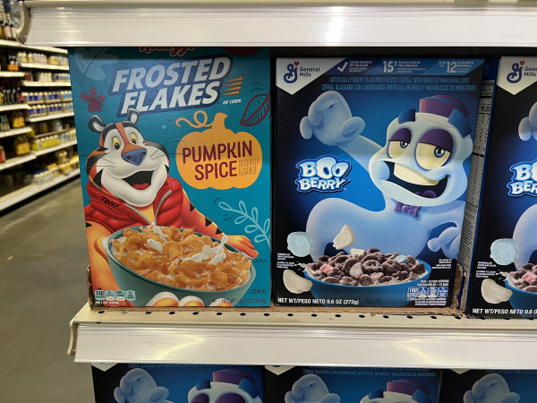 Two cereal boxes. 
On the left is a Kellogg’s “Pumpkin Spice” Frosted Flakes box with To y the Tiger in a winter vest. 
In the right is a box of General Mills Boo Berry cereal with the cartoon mascot. 