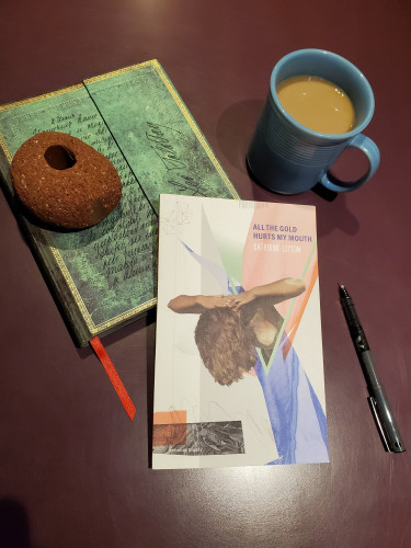 The poetry collection All the Gold Hurts My Mouth by Katherine Leyton (Goose Lane Editions), with its intriguing collage cover, sits with a notebook with an ornate green cover, a piece of red brick, a black pen and coffee in a blue cup
