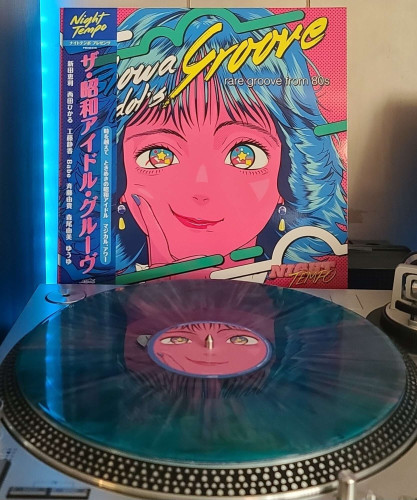 A Transparent Green with Pink and Blue Splatter vinyl record sits on a turntable. Behind the turntable, a vinyl album outer sleeve is displayed. The front cover shows artwork of a Japanese style idol from the 80s holding her finger to her cheek in a cute manner