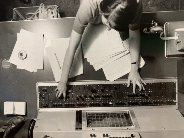 An overhead shot with Jean Hall having both hands on control buttons on the control panel of the ADVIDAC Computer at Argonne National Laboratory in the middle to late 1950s.  There are papers on the table, a paper tape machine, a paper tape bin below. Landscape orientation black and white photo is of the panel, and Hall w/ the photo perspective from directly above.


