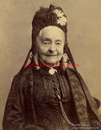 This is a photo of Margaret Ann Neve.

It is a sepia-coloured photo from the Victoria age. 

It shows an elderly lady looking straight into the camera.
She wears black Victorian garb, with a high collar, many small buttons, and a pocket watch on a silver chain. 

Her hair is in two tight ringlets extending from her ears, clearly white, and her head is topped with a feathered hat, sporting two white flowers.

Her face is aged but pleasant. She has hooded sharp eyes, an elegant nose, and a small smile grazes her thin lips. Her right eyebrow is slightly tilted upward as if to say that she is not done with mischief yet.

The background is blank.
