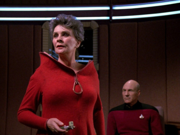 Admiral Satie wearing a red high color dress with Picard sitting in the background 