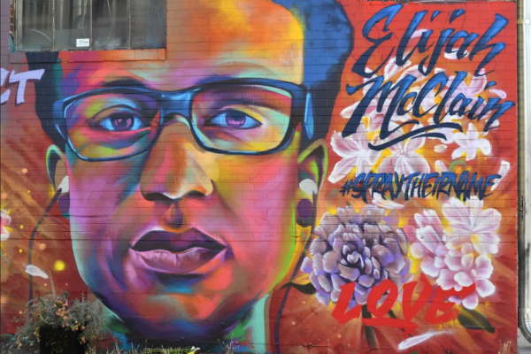 A mural of Elijah McClain is painted on the wall in his memory. it shows the 23-year-old massage therapist with earbuds in his ears. The murals background in various shades of red and orange also show flowers and the words: "Elijah McClain – respect – #SprayTheirName".