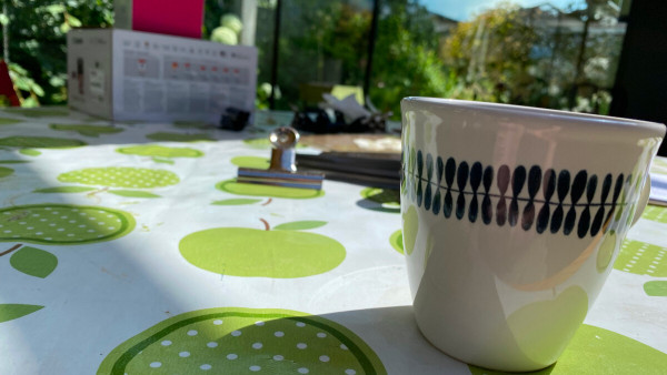 A close up of a table with a mug looming in the foreground, a tablecloth decorated with green apples and a box in the background.