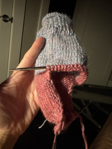 Hand holding a sock in a worsted yarn. Cuff and top of leg in a light blue yarn with speckles of other colors including pink. Cuff ribbed, leg stockinette. Color change to pink. Heel flap and turn completed. Flexiflip needles hanging.