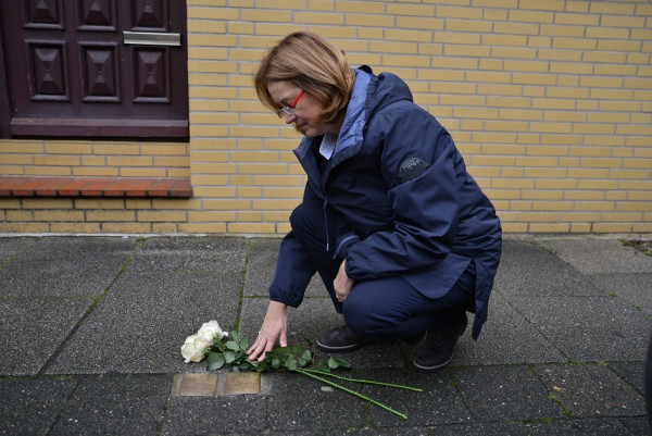 Bürgerschaftspräsidentin Antje Grotheer kniet vor den Stolpersteinen von Anna und Carl Stiegler und legt zwei weiße Rosen daneben. Sie trägt eine blaue Jacke und schaut ernst auf die Stolpersteine. Im Hintergrund ist eine gelbe Hauswand zu erkennen.
