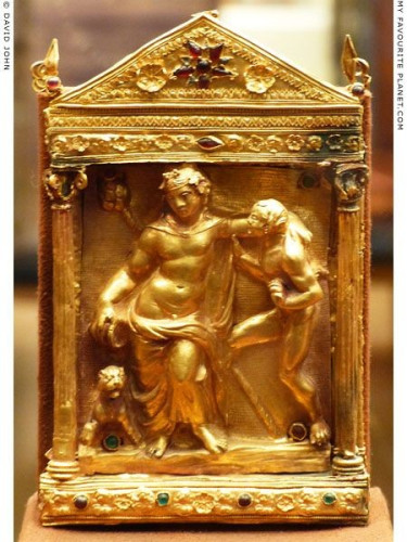 Golden naiskos relief of Dionysos framed by two Corinthian columns and a triangular pediment. The god is drunk, leaning on a satyr friend for support. To his feet, a panther crouches. Dionysos is depicted with a cloak pinned above his breast and a crown in his long, lush hair adorned with clusters of grapes. He personifies the abundance of a good life through his soft, sensual body.