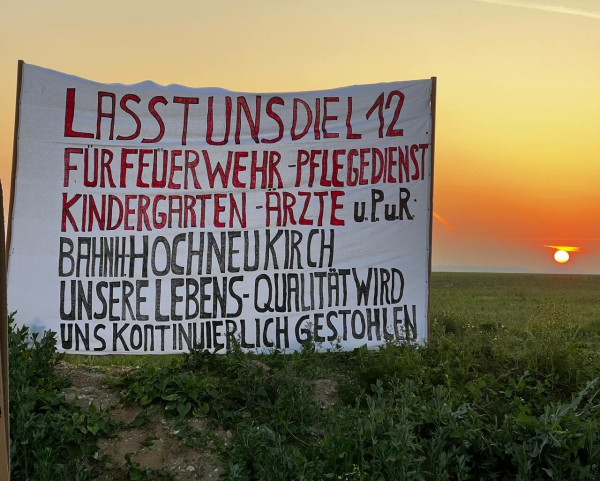 Banner am Wall des Tagebau Garzweiler bei Sonnenaufgang, mit der Aufschrift

"Lasst uns die L12
für Feuerwehr - Pflegedienst
Kindergarten - Ärzte u. P.u.R
Bahnh. Hochneukirch
Unsere Lebensqualität wird
uns kontinuierlich gestohlen".