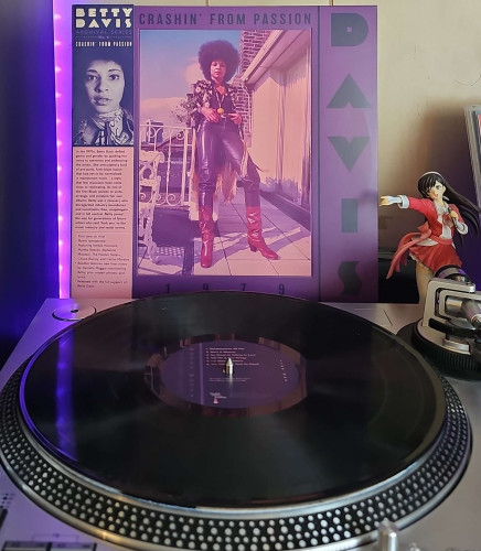 A black vinyl record sits on a turntable. Behind the turntable, a vinyl album outer sleeve is displayed. The front cover shows Betty Davis posing against a railing outside a home.

To the right of the album cover is an anime figure of Yuki Morikawa singing in to a microphone and holding her arm out. 