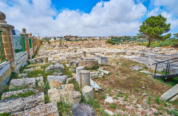 Rocky, shrubby landscape thickly littered with pieces of ancient columns and remains of other stone structures. The sky is blue with bright white clouds.