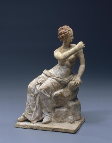 A clay figurine of Aphrodite seated on a rock. She is turning to her left, suggesting this might have originally been a sculpture group. She is wearing her reddish-brown hair in a neat bun, a himation around her hips leaving her topless. She seems to have held something in her right hand, raised at shoulder height, but whatever it was got lost to time.