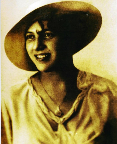 Élisabeth Le Port in sepis tinted black & white head and shoulders photo, wearing tilted widebrimmed hat and a pale dress with plaited detail at neck. dark hair curl on forehead, smiling looking photo left