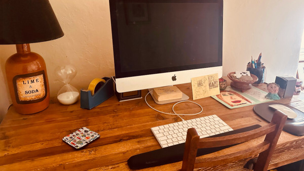 A computer on a writer's desk, with sketches, a clay sculpture and a pot of pens to the side.