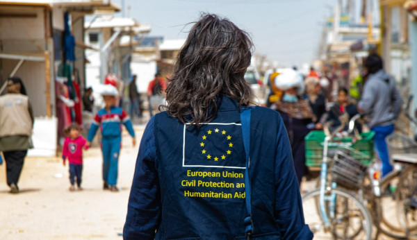 A person with a blue jacket with a yellow text on it. 

The text says: "European Union Civil Protection and Humanitarian Aid". 

Above the text, there is a EU Emblem.

The person has long curly hair and is seen from behind. 

In the background, a street full of people. 