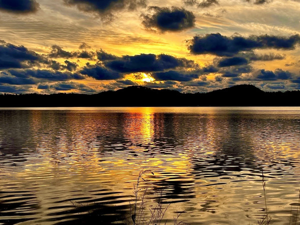 The sun peeks through clouds casting a golden light in the sky and reflected off a lake. In the center, it shines through an oval opening with a dark cloud arching overhead like a brow.