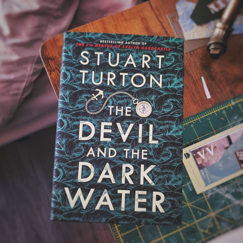 A copy of Stuart Turton's book, The Devil and the Dark Water, on a wooden desk, beside a green cutting mat.
