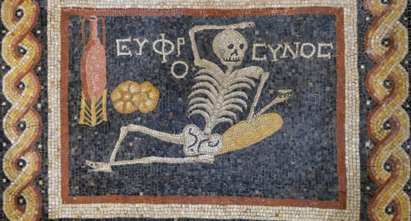 Section from a C3rd BCE mosaic. This panel has a dark blue background with a red and white panel border. The scene shows a skeleton holding a dish while reclining while next to them are two loaves of bread and an amphora of wine resting in a wooden stand.