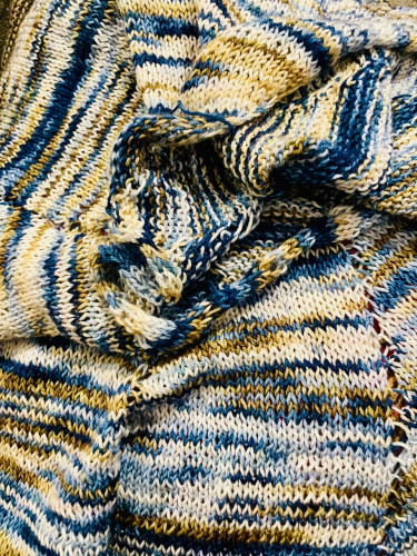 A hand knit scarf in colors of ivory, moss green, brown, and shades of light blue to dark blue. The pattern and the striping colors appear in a sort of swirl of stockinette stitch.