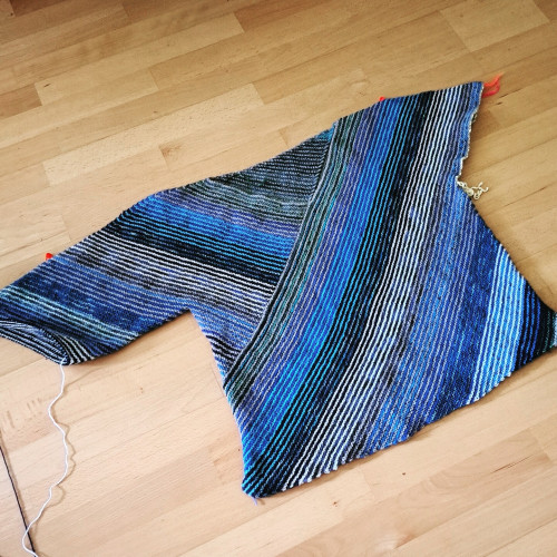 Unfininished hand knitted sweater in different shades of blue, green and grey leider out flat on a wooden floor.