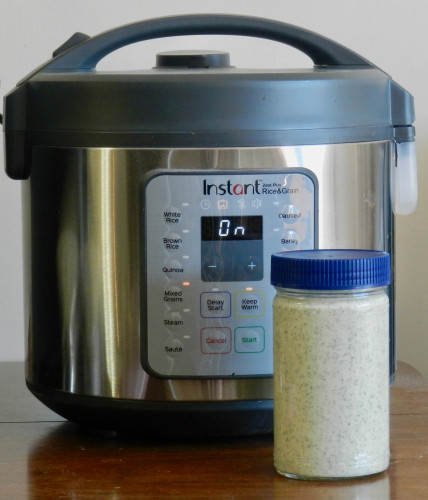 An Instant Zest grain cooker with the control panel showing "On" and "Mixed Grains." In front is a cylindrical glass jar with a blue cap filled with a light-green liquid.
