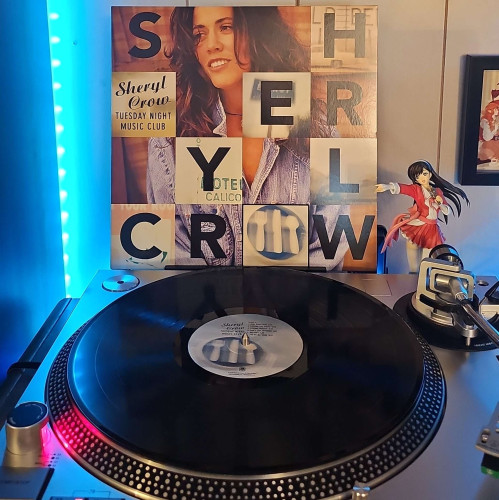 A black vinyl record sits on a turntable. Behind the turntable, a vinyl album outer sleeve is displayed. The front cover shows Sheryl Crow in a denim shirt. Her name is spelled out in blocks in which some show images of signs. 