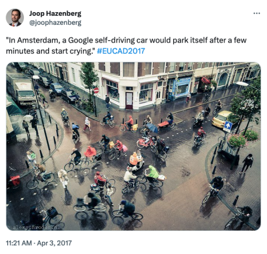 Photo of a busy intersection in Amsterdam, from 45 deg above. There are a lot of cyclists, pedestrians and a few parked cars.