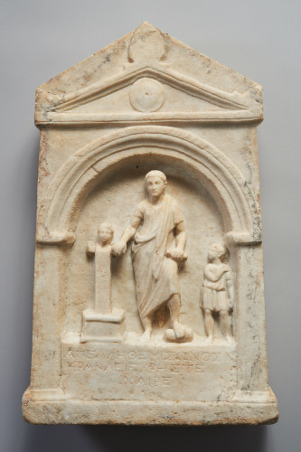 A funerary stele with a scene of a man with a youth beside him. He holds a cylinder (scroll?) in his left hand and seems to rest his left foot on a cylinder as well. His right hand rests on a herm. Below is an inscription in Greek.
