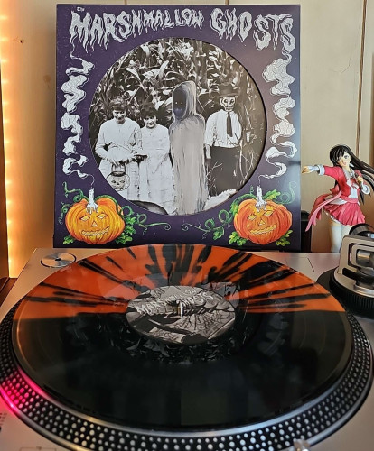 A Orange And Black Split w/ Black Splatter vinyl record sits on a turntable. Behind the turntable, a vinyl album outer sleeve is displayed. The front cover shows pumpkin candles and a cutout that shows creepy people in a cornfield. 

To the right of the album cover is an anime figure of Yuki Morikawa singing in to a microphone and holding her arm out. 
