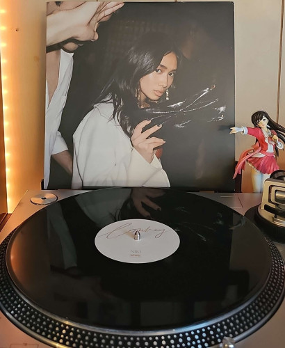 A black vinyl record sits on a turntable. Behind the turntable, a vinyl album outer sleeve is displayed. The front cover shows NIKI looking at the camera and holding her purse up. Next to her a man is covering his face. 

To the right of the album cover is an anime figure of Yuki Morikawa singing in to a microphone and holding her arm out. 