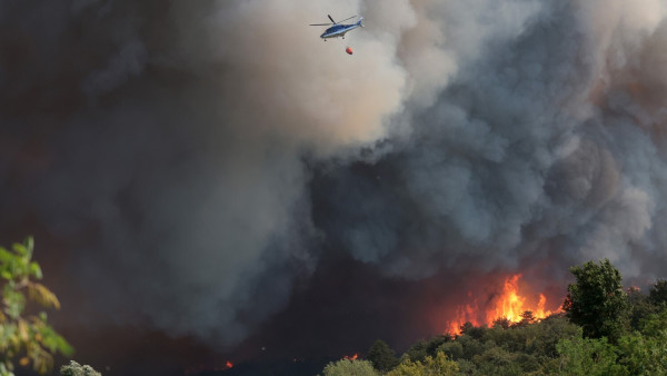 A firefighting helicopter drops water on a fire, whose dark smoke covers almost all the area.
