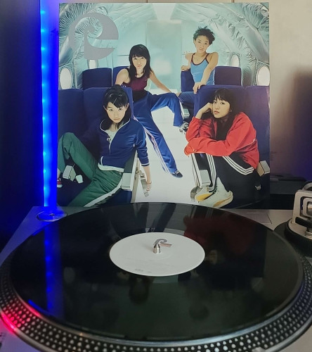 A black vinyl record sits on a turntable. Behind the turntable, a vinyl album outer sleeve is displayed. The front cover shows the 4 members of Speed sitting in various position in airline-style seats