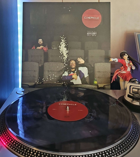 A Clear w/Black Smoke vinyl record sits on a turntable. Behind the turntable, a vinyl album outer sleeve is displayed. The front cover shows Sirius Blck acting surprised in a movie theater. His popcorn is flying straight up. There is another guy sitting a couple rows behind him. 

To the right of the album cover is an anime figure of Yuki Morikawa singing in to a microphone and holding her arm out. 