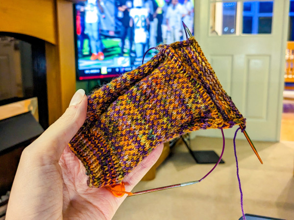 Holding up an in-progress knitted sock, in a slip stitch pattern of orange and purple. The leg is complete and the heel flap is just starting to come into view. In the background, the Miami Dolphins football game is on TV.