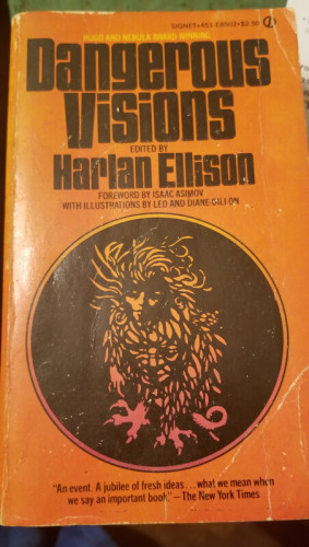 A beat-up paperback book called Dangerous Visions edited by Harlan Ellison. It's orange and has a circular black coin or disc on the cover with some kind of mythological creature on it that has claws, feathers, and a human face. 