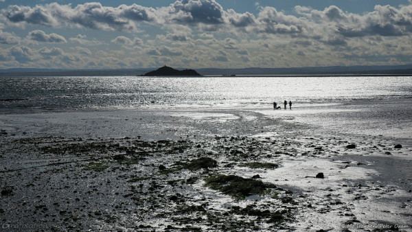 A seascape, with wet sand and water reflecting the sunlight. The sky is blue and has fluffy clouds. In the middle distance is the silhouette of a pointed island with a tall structure at its top. Close to the centre of the photo are the silhouettes of three people and two dogs walking on the wet beach. The foreground is low rocks and seaweed. In the distance is the opposite shore of the bay, an indistinct line of hills cloaked in mist which is actually the northern coastline of Edinburgh. Most of the shot is filled with reflected sunlight coming from almost directly overhead.