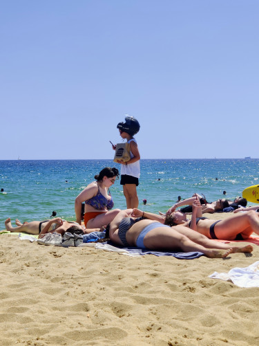 A delivery worker on the beach with a motorcycle helmet perched on her head, and with a smartphone in one hand and a brown Burger King bag in the other. Presumably, she's trying to find whomever placed the order. Some sunbathers are in the foreground and the Mediterranean sea is in the background.