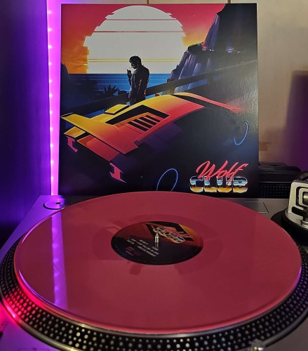 A pink vinyl record sits on a turntable. Behind the turntable, a vinyl album outer sleeve is displayed. The front cover shows digital artwork of a man standing next to a Lamborghini near a railing on a mountain road lighting a cigarette. In the distance, cop cars are coming around a bend in the road. In the background is a sunset and water