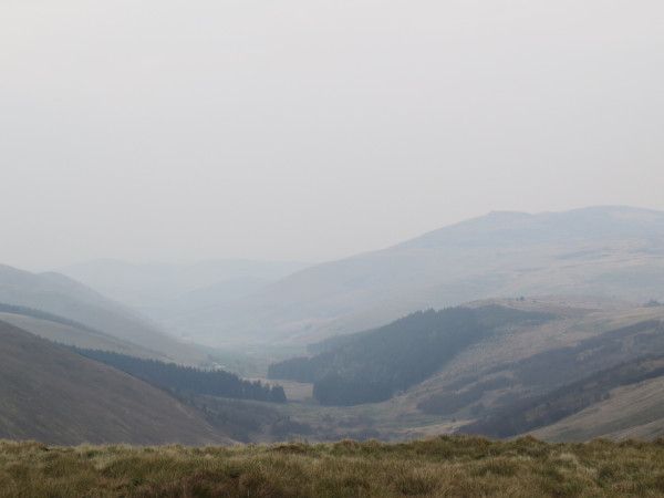 A misty image of Northumbrian moorland, greenish-brown grass and heather in the foreground dropping into a deep valley, beyond which layers of hills fade into the horizon.
