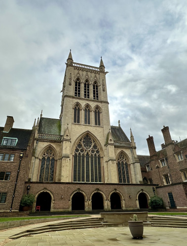 Photo of the Chapel of St John’s College, Cambridge, taken from within the College grounds—specifically the chapel court. A large, Neo-Gothic structure in yellow stone, it soars above the adjacent brick buildings, piercing the dull, early-December sky above. 