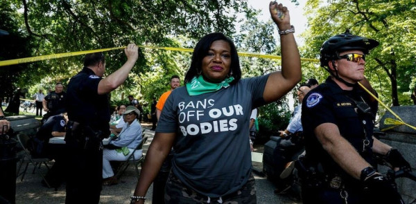 With police behind her, a woman wearing a T-shirt that reads “Bans off our bodies,” holds her left fist in the air.