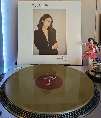 A Metallic Gold vinyl record sits on a turntable. Behind the turntable, a vinyl album outer sleeve is displayed. The front cover shows Laufey leaning against a wall, holding her left arm with her hand. She is looking off camera.. 

To the right of the album cover is an anime figure of Yuki Morikawa singing in to a microphone and holding her arm out. 
