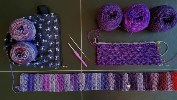 On a black tile surface the following items are displayed, going clockwise starting top right: 
- three cakes of purple yarn, all three slightly different in hue
- a knitted rectangle on the needles, in the three purple yarns using garter stitch showing rainbow colored stitch markers
- a small crocheted scarf in progress in a selfstriping yarn that has different reds and purples with grey and lavender in between 
- a black project bag with purple wizards on the fabric with two yarn cakes on top in the selfstriping colorway of the scarf
