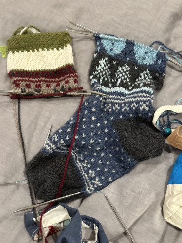 Left: cuff of an Xmas stocking in green, cream, and maroon with some black stitches top of dog head. Right: most of an Xmas stocking in shades of blue and white/ivory. Middle pattern is pine trees. Top of pattern is bottom half of snowmen bodies.