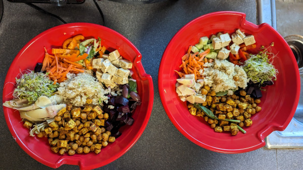 Two red bowls containing a giant Greek salad: greens and baby spinach, beets, chickpeas, sweet potato, artichoke hearts, quinoa, carrots