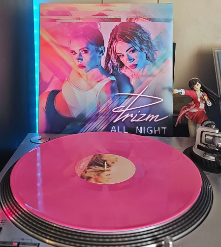 A pink vinyl record sits on a turntable. Behind the turntable, a vinyl album outer sleeve is displayed. The front cover shows the two members of Prizim. 

To the right of the album cover is an anime figure of Yuki Morikawa singing in to a microphone and holding her arm out. 