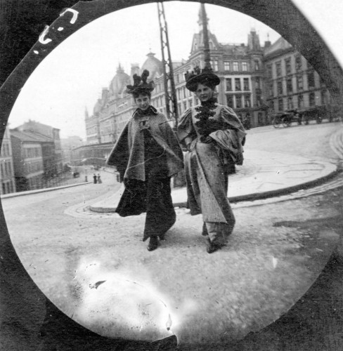 A black and white photo showing two Victorian women walking on a street. The photo has some distortion from being taken on a concealed camera
