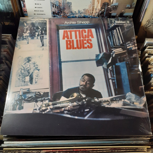 Album cover features a photograph of Archie Shepp seated at a piano, with a cigarette in his mouth and window behind him.  A saxophone lies on top of the piano.  A shelf with messy stacks of books is seen to the side, along with a couple of green beer bottles, and a poster of two black Olympic athletes raising their fists in protest on the podium at the 1968 Olympic Games.